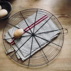 May include: A black wire cooling rack with a white and red striped kitchen towel. An egg rests on the towel, and two more eggs are in a dark bowl. The items are on a light wood surface.