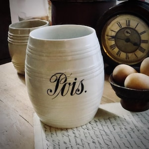 May include: Cream-colored ceramic jar with the word "Pois" in black script. The jar has horizontal ridges and is next to a similar jar, a clock, and a small bowl with eggs. The background includes a vintage aesthetic.