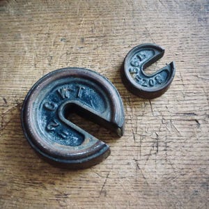 May include: Two antique, cast iron weights with a dark patina sit on a wooden surface. The larger weight is a partial circle with the number "2" and other markings. The smaller weight is also a partial circle with the number "208" and other markings.