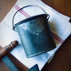 May include: An antique metal milk pail with a handle, the words "EXPRESS DAIRY CO" are embossed on the side. The pail sits on a white cloth with red stripes, alongside a wooden-handled tool and a metal blade.