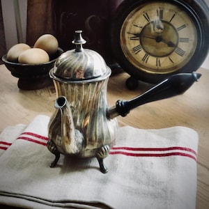 May include: A vintage silver coffee pot with a black handle sits on a white and red striped cloth. A dark bowl of eggs and a classic clock are in the background. The coffee pot has a decorative, ridged design.