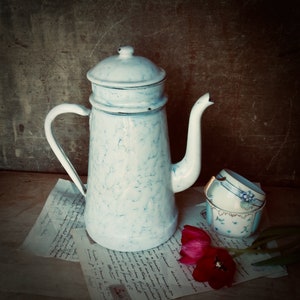 May include: A white enamel coffee pot with a blue marbled design and a blue and white porcelain teacup with gold trim. The coffee pot has a spout and a handle. The teacup is sitting on a piece of paper with writing on it.