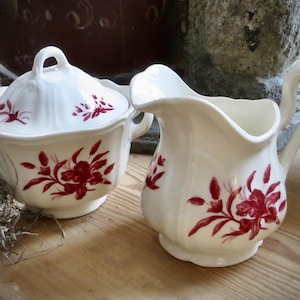 May include: A white ceramic sugar bowl with a lid and a creamer pitcher, both adorned with a red floral pattern. The sugar bowl has two handles and a lid with a small handle. The creamer has a curved handle and a spout.