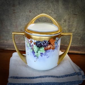 May include: A white porcelain sugar bowl with a gold lid and handle. The bowl is decorated with a hand-painted design of purple grapes, green leaves, and brown vines.