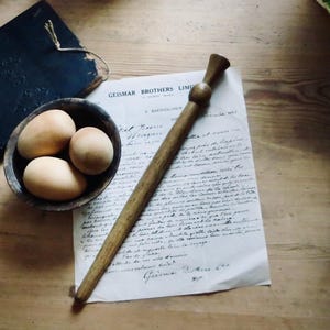 May include: A wooden rolling pin and a bowl of three eggs sit on a wooden surface next to a piece of paper with handwritten text. A dark blue book is in the background. The paper has the text "Geismar Brothers Limited" printed on it.