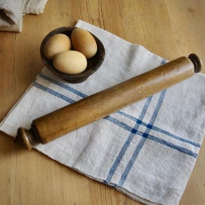 May include: A cylindrical wooden rolling pin with rounded handles at each end, positioned diagonally across a white linen cloth featuring blue stripes, accompanied by a small dark wooden bowl containing three smooth, light brown eggs.