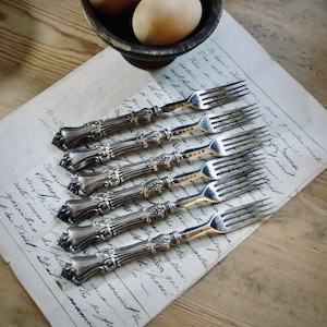 May include: Six antique silver forks with ornate handles are arranged on a piece of aged paper with handwritten script. The forks have a detailed design and are placed on a wooden surface. A small bowl with eggs is in the background.