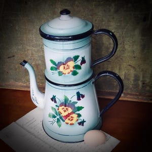May include: A vintage blue and white enamel coffee pot with a floral design. The pot has a black rim and handle. The flowers are yellow, purple, and green. The pot is sitting on a piece of paper with writing on it.