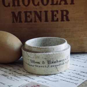 May include: A small, off-white ceramic jar with the text "Allen & Hanburys, 1 Vere Street, Cavendish Sq" is next to a brown, egg-shaped object. The jar has a cracked, aged appearance. The background includes a wooden box with the text "CHOCOLAT MENIER".