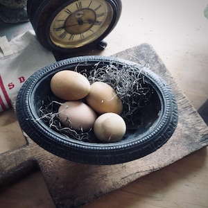 May include: An antique black oval bowl filled with four brown eggs and dried grass, resting on a wooden cutting board. A vintage clock and a canvas bag with red stripes are in the background, creating a rustic, farmhouse aesthetic.