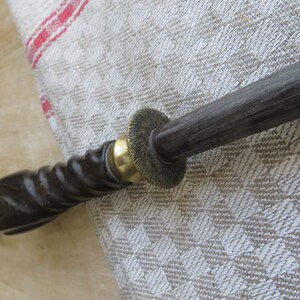 May include: A close-up of a dark-colored knife sharpener with a wooden handle. The handle has a spiral design and is accented with brass-colored metal. The sharpening rod is dark gray.