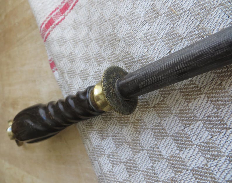 May include: A close-up of a dark-colored knife sharpener with a wooden handle. The handle has a spiral design and is accented with brass-colored metal. The sharpening rod is dark gray.