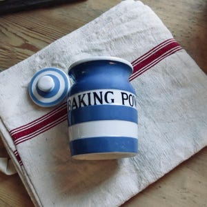 May include: A blue and white ceramic baking powder jar with a matching lid. The jar has a white stripe and the words "BAKING POW" in white. It sits on a white linen towel with red stripes.