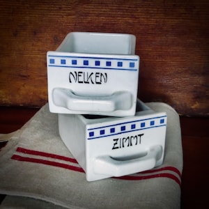 May include: Two white ceramic spice jars with blue and white trim. The top jar says "NELKEN" and the bottom jar says "ZIMMT".