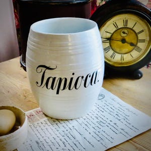 May include: A white ceramic tapioca jar with black script lettering. The jar has a barrel shape with horizontal ridges. An antique clock and a handwritten document are in the background.