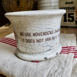 May include: A white ceramic shaving mug with a prominent crackle glaze finish. The mug features dark brown printed text that reads: WE USE HOVENDENS SHAVING SOAP IT DOES NOT IRRITATE THE SKIN. The base is flared and shows minor dark wear marks.