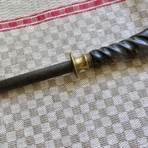 May include: A dark, wooden knife sharpener with a brass bolster and a twisted, dark handle. The sharpener is set against a patterned, neutral-toned fabric with a red stripe.