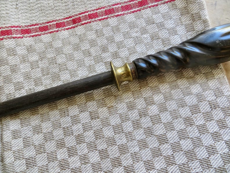May include: A dark, wooden knife sharpener with a brass bolster and a twisted, dark handle. The sharpener is set against a patterned, neutral-toned fabric with a red stripe.