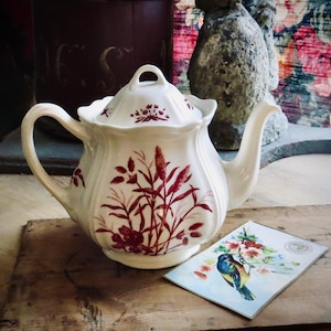 May include: A white ceramic teapot with a floral design in burgundy, featuring a curved handle and a lid. A postcard with a bird illustration rests beside it on a wooden surface. The teapot has a capacity of approximately 40 ounces.