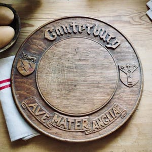 May include: A circular wooden serving board featuring the carved text Canterbury at the top and Ave Mater Angliae at the bottom. The board includes two carved heraldic shields on the sides, one depicting a lion and the other a cross symbol.