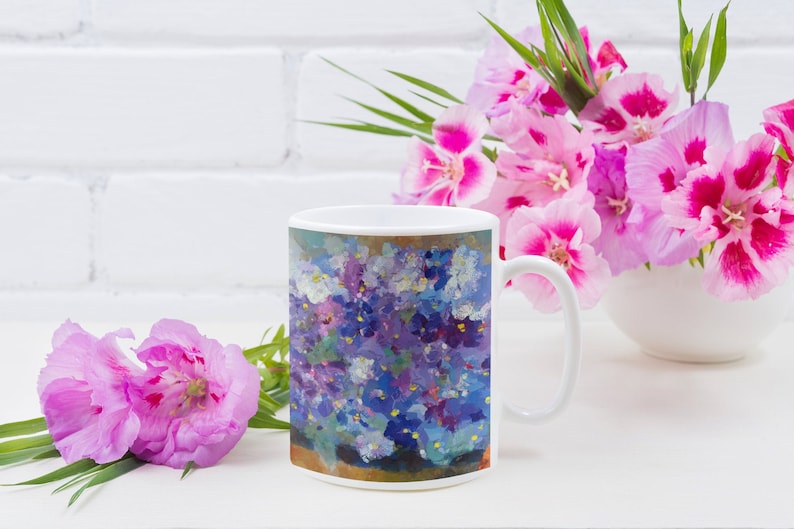 Pu&ograve; includere: Tazza in ceramica bianca con un design floreale colorato con fiori viola, blu e bianchi. La tazza ha un manico ed &egrave; appoggiata su una superficie bianca.