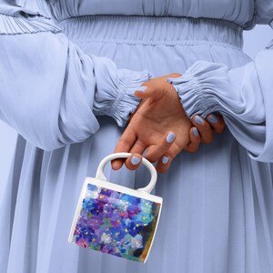 Pu&ograve; includere: Una tazza in ceramica bianca con un design floreale colorato con fiori viola, blu e gialli. La tazza &egrave; tenuta da una mano dietro una persona che indossa un vestito blu chiaro.