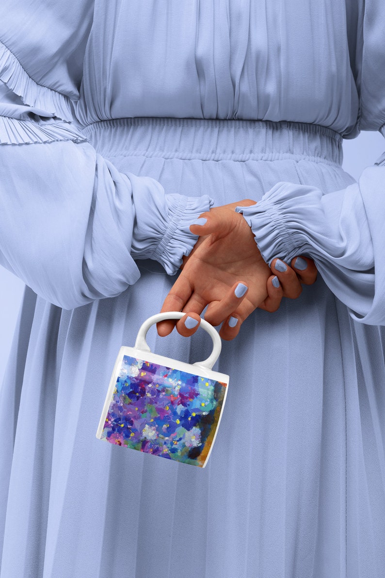 Pu&ograve; includere: Una tazza in ceramica bianca con un design floreale colorato con fiori viola, blu e gialli. La tazza &egrave; tenuta da una mano dietro una persona che indossa un vestito blu chiaro.