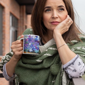 Pu&ograve; includere: Una donna tiene in mano una tazza di ceramica bianca con un motivo floreale. La tazza presenta un bouquet di fiori viola e blu con accenti bianchi. La donna indossa uno scialle a motivi verdi e bianchi e guarda da un'altra parte.