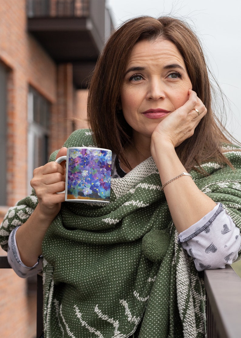 Pu&ograve; includere: Una donna tiene in mano una tazza di ceramica bianca con un motivo floreale. La tazza presenta un bouquet di fiori viola e blu con accenti bianchi. La donna indossa uno scialle a motivi verdi e bianchi e guarda da un'altra parte.