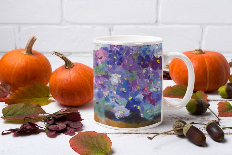 Pu&ograve; includere: Una tazza in ceramica bianca con un design floreale colorato con fiori viola, blu e bianchi. La tazza &egrave; appoggiata su una superficie bianca con foglie autunnali e zucche.