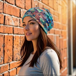 May include: A slouchy beanie hat featuring an abstract, painterly pattern of blue, teal, purple, and gold heart shapes. The fabric has a soft, blended appearance with varying shades of cool tones and warm metallic accents throughout the design.