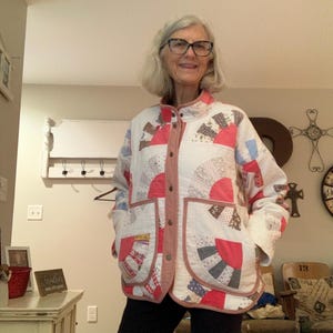 May include: A quilted jacket featuring a white base with a repeating fan-shaped pattern in shades of red, grey, and patterned fabric. The jacket has a stand-up collar, a front button closure, and two large patch pockets with a solid red trim.