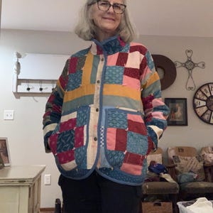 May include: A colorful patchwork jacket with a stand-up collar and button closure. The jacket features a variety of fabric squares in shades of pink, blue, yellow, and red. The jacket has two pockets and is trimmed with blue fabric.