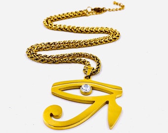 Könnte beinhalten: Eine goldene Kettenkette mit einem goldenen Anhänger in Form eines Auges mit einem klaren Kristall in der Mitte. Der Anhänger ist eine stilisierte Darstellung des Horusauges, ein altes ägyptisches Symbol für Schutz und Gesundheit.