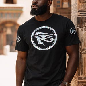 May include: Black t-shirt featuring a circular graphic with an Eye of Horus design. The shirt has short sleeves and a crew neck. A similar graphic is on the left sleeve. The person is wearing a gold watch and khaki pants.