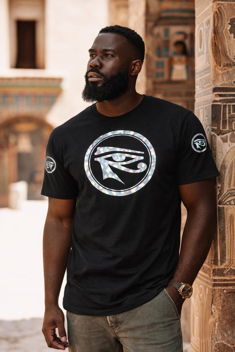 May include: Black t-shirt featuring a circular graphic with an Eye of Horus design. The shirt has short sleeves and a crew neck. A similar graphic is on the left sleeve. The person is wearing a gold watch and khaki pants.