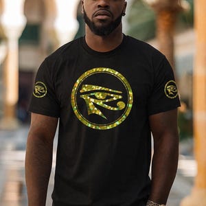 May include: Black t-shirt featuring a gold-colored Eye of Horus design in a circular frame on the front. The short-sleeved shirt also has a smaller Eye of Horus emblem on the left sleeve. The person is wearing a watch with a leopard print band.