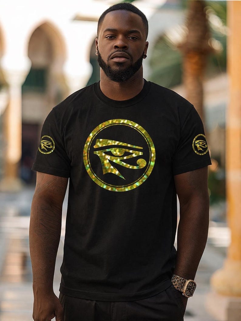 May include: Black t-shirt featuring a gold-colored Eye of Horus design in a circular frame on the front. The short-sleeved shirt also has a smaller Eye of Horus emblem on the left sleeve. The person is wearing a watch with a leopard print band.