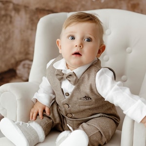 Puede incluir: Un bebé varón con un chaleco marrón y una corbata de lazo, una camisa blanca y zapatos grises está sentado en una silla blanca. Está mirando hacia arriba y tiene una expresión de sorpresa en su rostro.