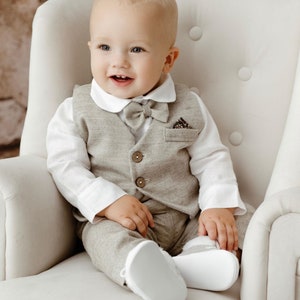 Può includere: Un bambino in un completo grigio chiaro, camicia bianca e papillon è seduto in una poltrona bianca. Il completo comprende gilet, pantaloni e pochette abbinata. Il bambino indossa scarpe bianche e sorride.