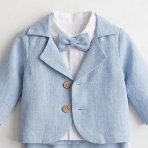Puede incluir: Una chaqueta azul claro de manga larga con solapa de muesca, que presenta dos botones redondos de madera en la parte delantera. Debajo lleva una camisa blanca de cuello rígido con una pajarita azul claro a juego en el cuello.
