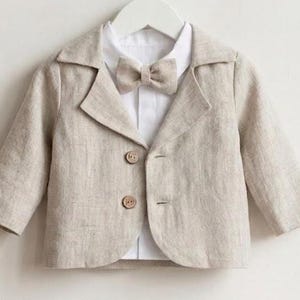 Puede incluir: Una americana de color beige con mangas largas y dos botones marrones, superpuesta sobre una camisa blanca de cuello con una pajarita beige a juego. La americana presenta una solapa de muesca y se muestra en una percha blanca de tela texturizada.