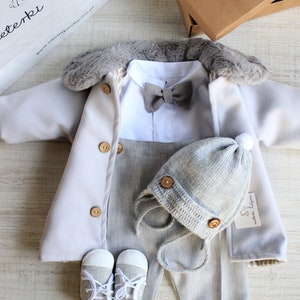 Könnte beinhalten: Ein Baby-Outfit-Set in Grau und Weiß. Das Set beinhaltet einen langärmeligen Mantel mit Kunstfellkragen, ein weißes Hemd mit Fliege, graue Hosen, eine graue Strickmütze und ein Paar grau-weiße Schuhe. Das Outfit liegt auf einer Holzoberfläche.