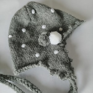 Può includere: Un berretto lavorato a maglia di colore grigio con piccoli pon pon bianchi sparsi sulla superficie. Il berretto presenta paraorecchie, un pon pon bianco più grande fissato sul lato e due lunghi lacci intrecciati sui bordi inferiori.