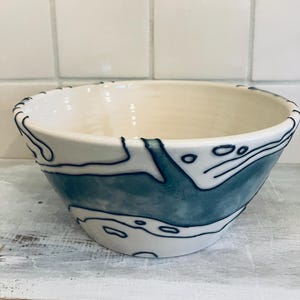 May include: A ceramic bowl with a cream-colored interior and a blue and white exterior design. The bowl features a blue watercolor-like pattern and black outline details. The rim has a black trim.