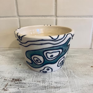 May include: A small, off-white ceramic bowl with a unique design. The bowl features a pattern of teal and navy blue organic shapes. The interior is a glossy off-white. The bowl sits on a white, distressed wooden surface.