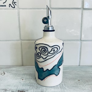 May include: A white ceramic oil bottle with a stainless steel spout. The bottle is decorated with a black and teal topographical design and a dotted red line. The bottle is designed for kitchen use.