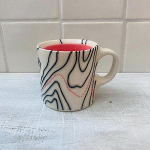 May include: A small, off-white ceramic mug with a black and pink topographic design. The interior of the mug is a vibrant red. The mug has a rounded handle and sits on a white surface.