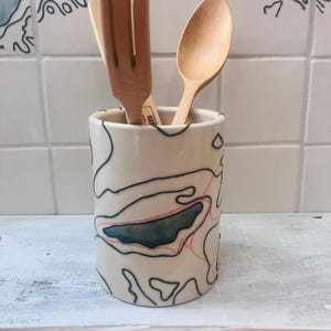 May include: A ceramic utensil holder with a cream-colored base, featuring a black and pink topographical map design. The holder contains a wooden spoon and a slotted spatula. The design wraps around the cylindrical shape.