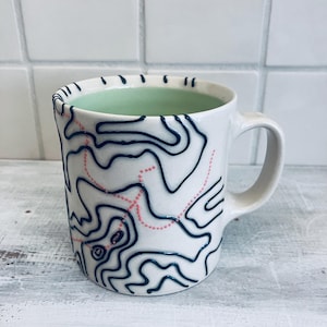 May include: White ceramic mug with a light green interior and a handle. The exterior features a topographical map design in dark blue and pink. The mug is a unique piece of drinkware.